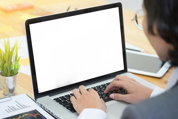Person typing on laptop with blank screen, surrounded by office items, depicting scary online moments trauma.