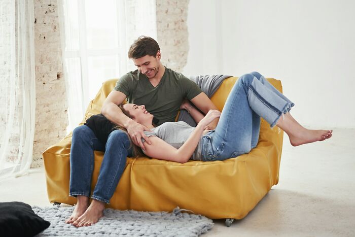Couple sharing a playful moment on a yellow couch, showing quirky affectionate behavior in a bright, cozy room.