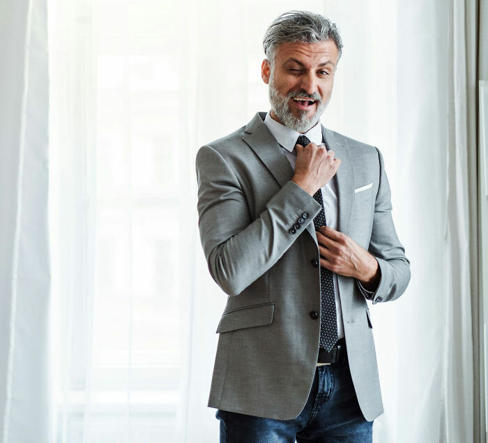 Man in gray blazer and polka dot tie winking while adjusting tie, representing male coworkers' bold comments at work.