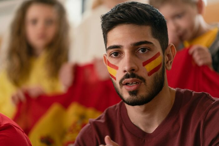 Young man with painted Spanish flag on cheeks, discussing big no no to do in your country, with people blurred in background.