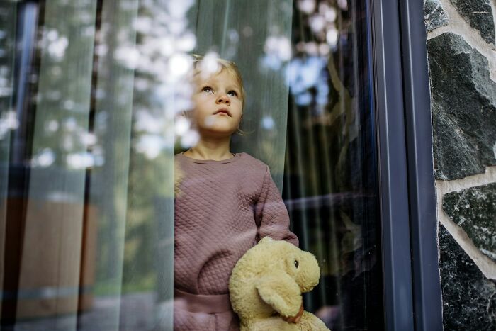 Young child holding a stuffed animal, looking sadly out a window reflecting trees, representing worst parents seen.