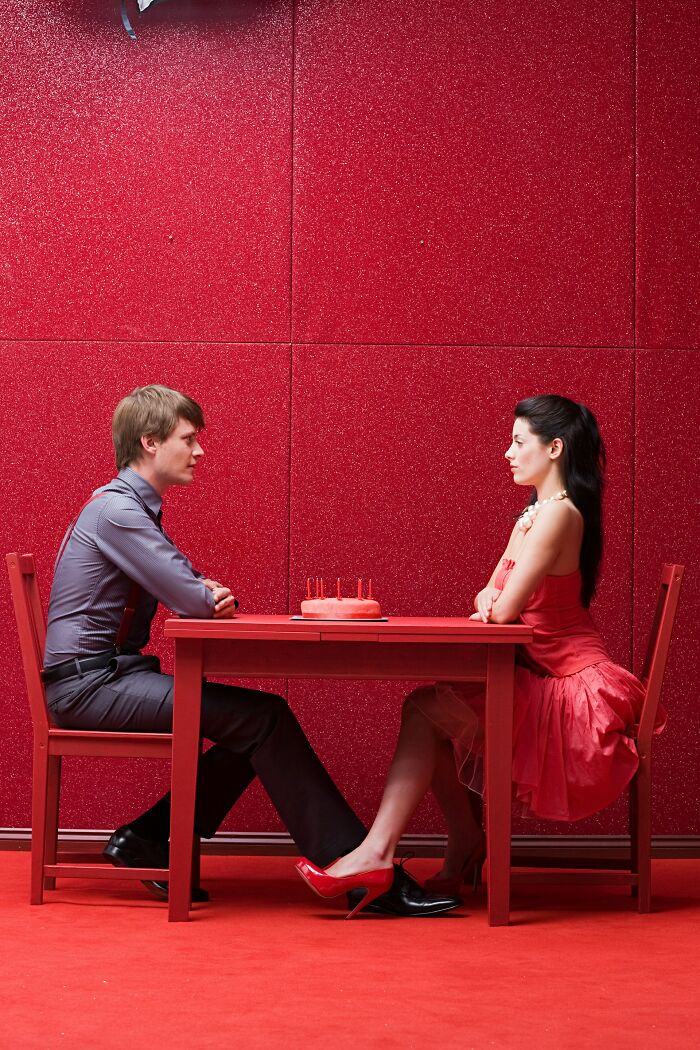 A tense couple sitting across a red table with a cake, illustrating challenges in modern dating experiences.