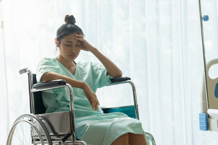 Pregnant woman in hospital gown sitting in wheelchair, holding head, reflecting pregnancy and postpartum challenges.