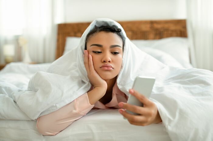 Young woman looking insecure while lying in bed under a blanket, holding a smartphone and feeling uncertain.