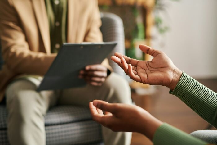 Therapist and client in a counseling session, therapist holding a clipboard listening while client gestures with hands.