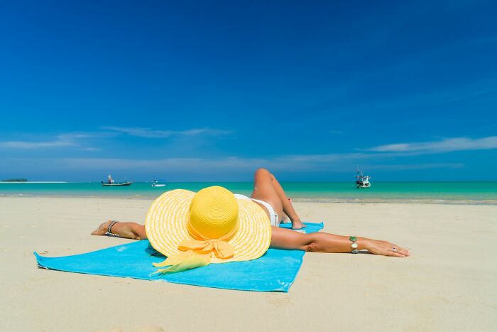 Woman wearing a yellow sunhat lying on a blue towel at the beach under clear skies, illustrating golden travel hacks.