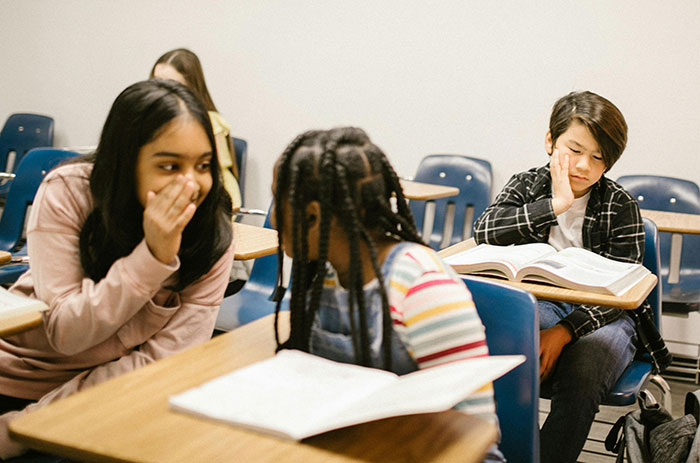 Two 5th grade girls whispering a revenge plot in classroom while a boy looks distressed sitting at his desk. Two 5th grade girls whispering a revenge plot in classroom while a boy looks distressed sitting at his desk.