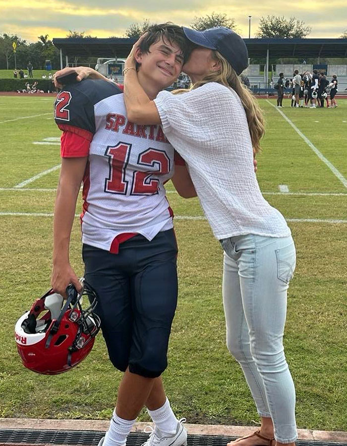 Teen football player wearing Spartans jersey embraces woman kissing his cheek on sports field.