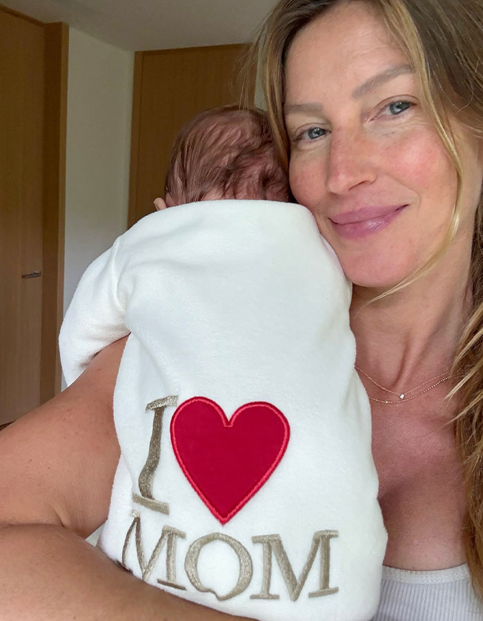 Gisele Bundchen holding her baby wearing a white outfit with I love mom text, facing away from the camera.