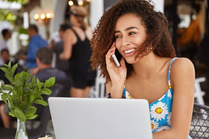 Smiling woman using phone and laptop at cafe, enjoying golden travel hacks to improve her trip planning experience.