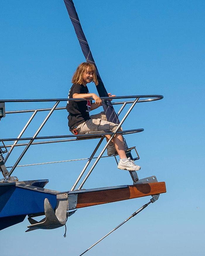 Singer sister of Greta Thunberg sitting on a sailboat railing, smiling against a clear blue sky background.