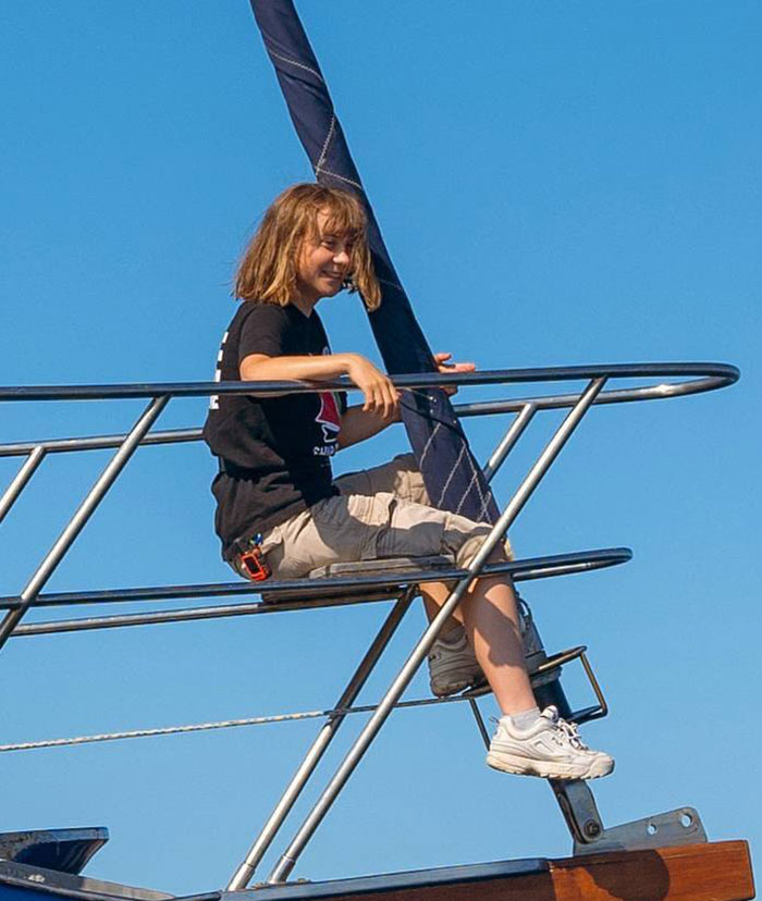 Greta Thunberg smiling and sitting on a boat railing under clear blue sky, linked to Chris Kebbo photographer rumors.