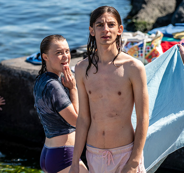 Greta Thunberg and Chris Kebbo at the water's edge, with Greta smiling and Chris standing shirtless holding a towel.