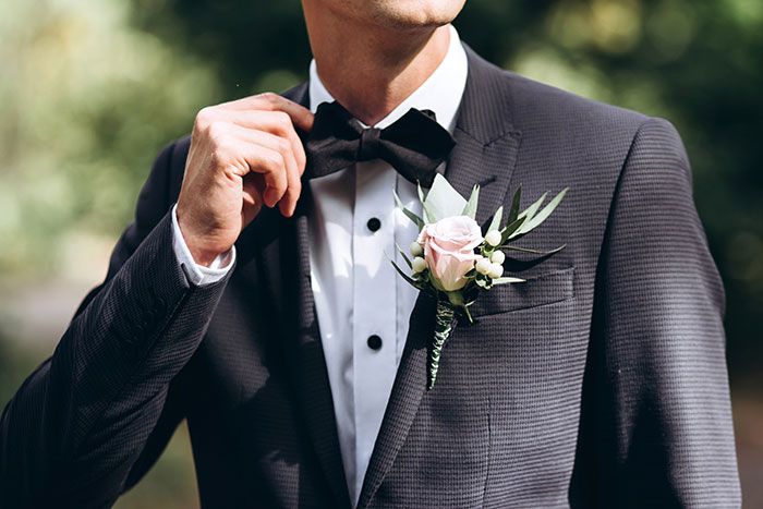 Man adjusting black bow tie wearing a dark suit with boutonniere at a Disneyland wedding ceremony outdoors.