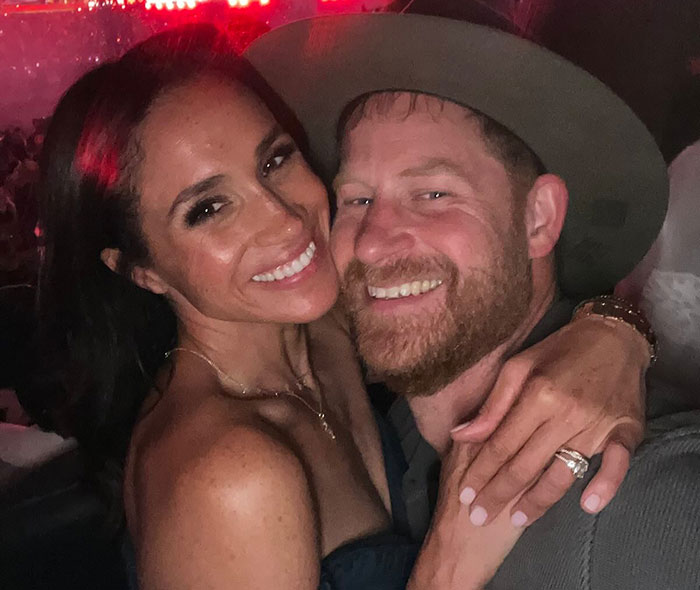 Meghan Markle and Prince Harry smiling and embracing at a nighttime event with warm lighting and casual attire.