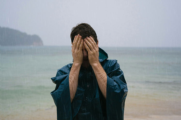Man in raincoat covering face in the rain on a tropical beach, illustrating realities of living in tropical places.