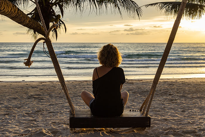 Person sitting on a swing under palm trees, looking at the ocean during sunset, capturing tropical living realities.