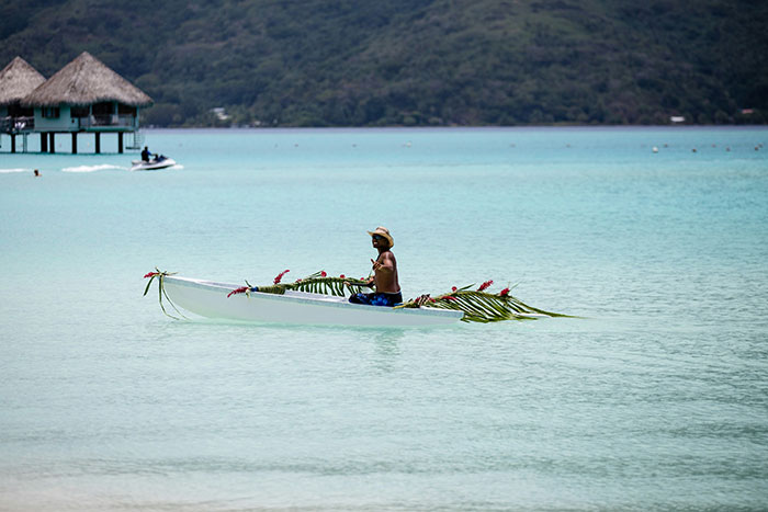 Man paddling a small decorated boat on clear turquoise water in a tropical place with overwater huts in the background