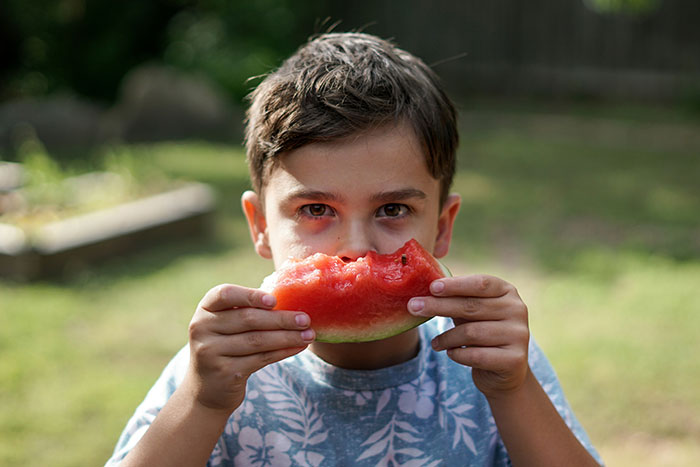 Young boy eating watermelon outdoors, capturing everyday realities of living in tropical places many dream of.