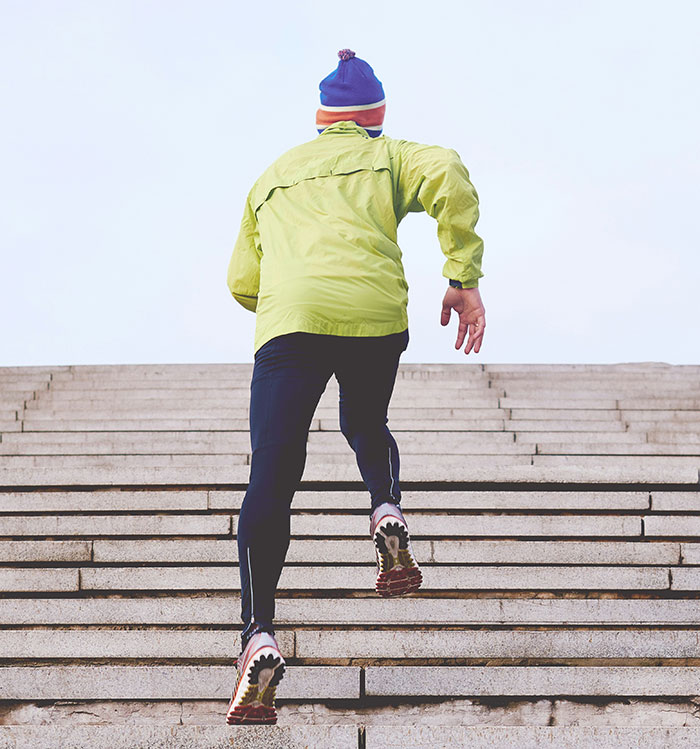 Person wearing a green jacket and blue hat running up stairs, symbolizing fitness and fat targeting in health and wellness.