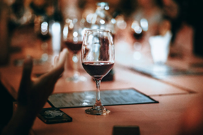 A glass of red wine on a table in a dimly lit setting with blurred background and people around.