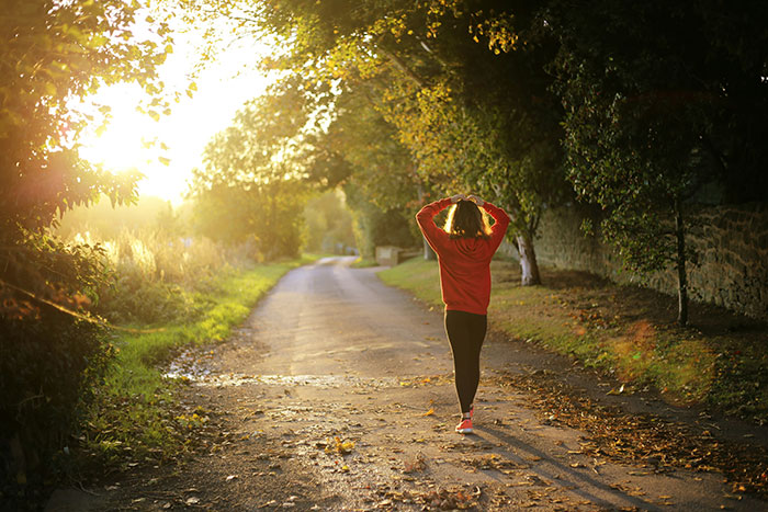 Person in red jacket walking on a sunlit path surrounded by trees, illustrating health and wellness outdoors.