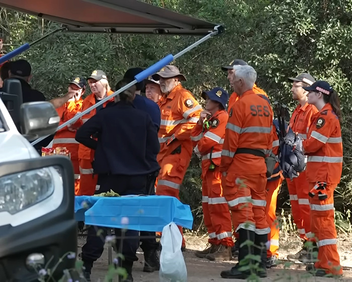 Group of SES volunteers in orange uniforms gathered outdoors near a vehicle during a funeral event with teen mourner present