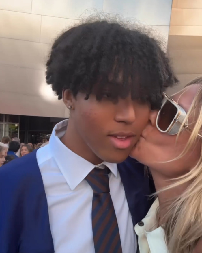 A woman wearing sunglasses kisses a young man dressed in a suit and tie at an outdoor event.