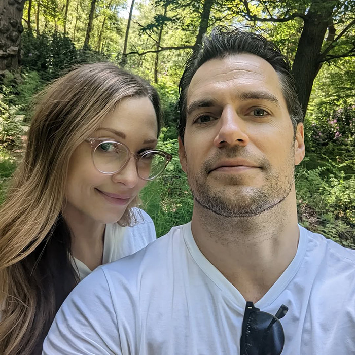 Henry Cavill outdoors with a new Highlander hairstyle, posing with a woman in a forest setting on a sunny day.