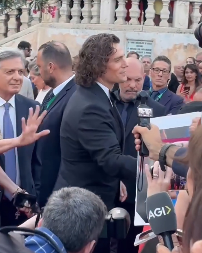Henry Cavill at a public event with curly Highlander hairstyle, interacting with fans and media amid a crowd.