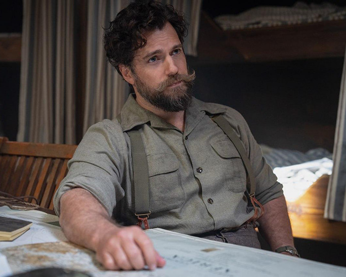 Henry Cavill with a new Highlander hairstyle, seated indoors wearing a grey shirt and suspenders, sporting a beard and curly hair.