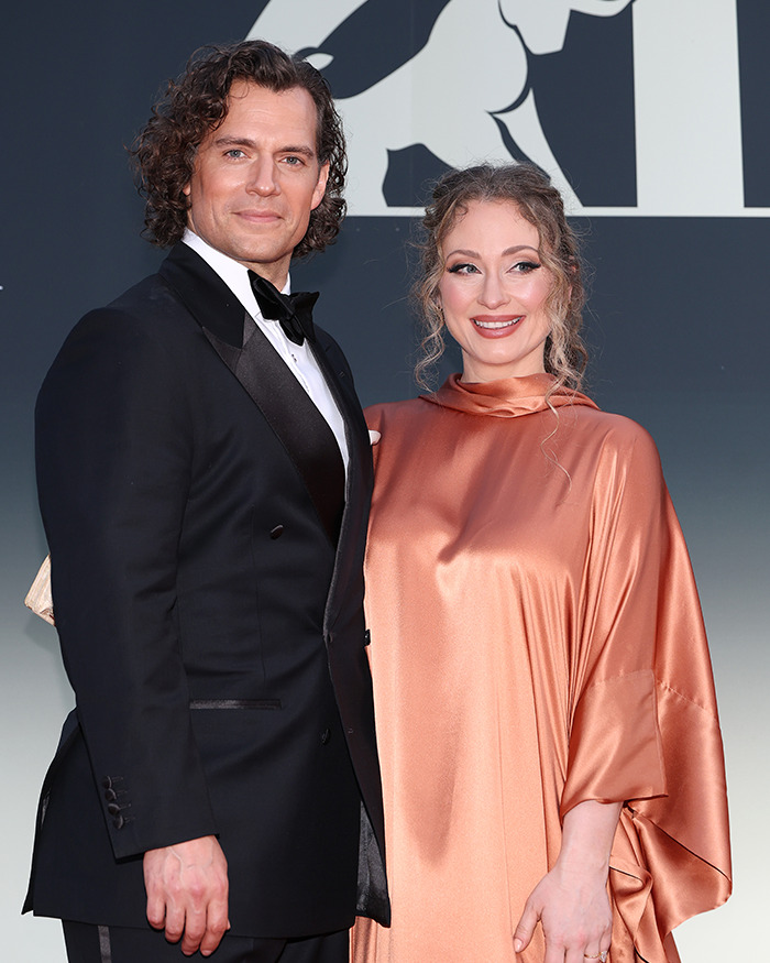Henry Cavill wearing a black tuxedo with a curly Highlander hairstyle posing with a woman in a shiny peach dress at an event.