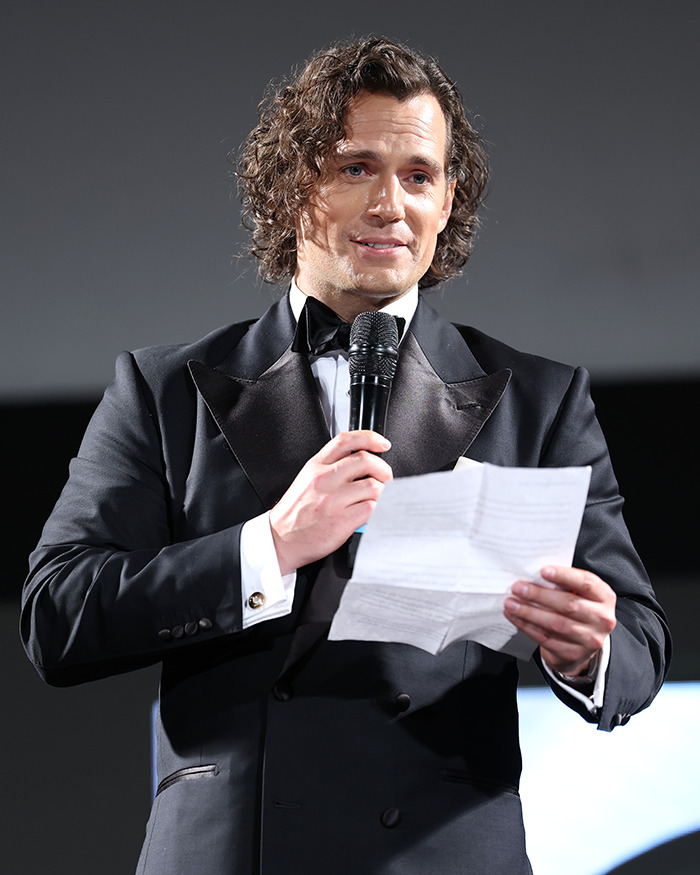 Henry Cavill with new Highlander hairstyle, wearing a black tuxedo and holding a microphone while reading from a paper.