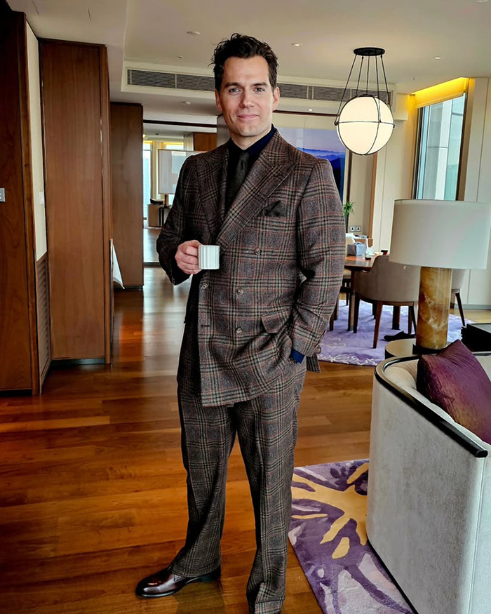 Henry Cavill in a brown plaid suit holding a cup indoors, showcasing his new Highlander hairstyle attracting fan attention.