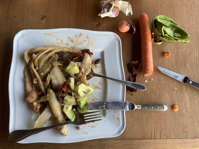 Leftover food scraps on a plate with utensils, alongside vegetable peelings and a knife on a wooden table showing poor people habits.