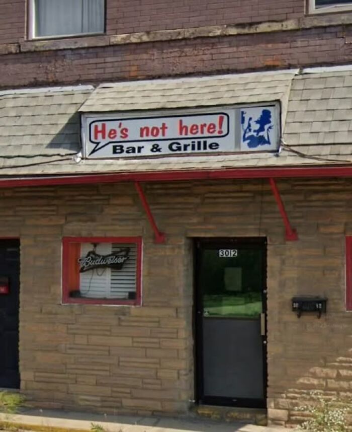 Humorous bar sign reading He's not here adding chaos and laughter to the world in a small building exterior.