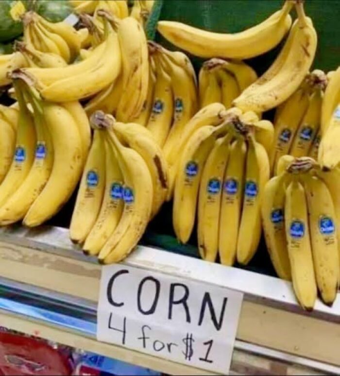 Bunches of bananas labeled as corn with a handwritten sign reading 4 for $1 in a chaotic grocery display.