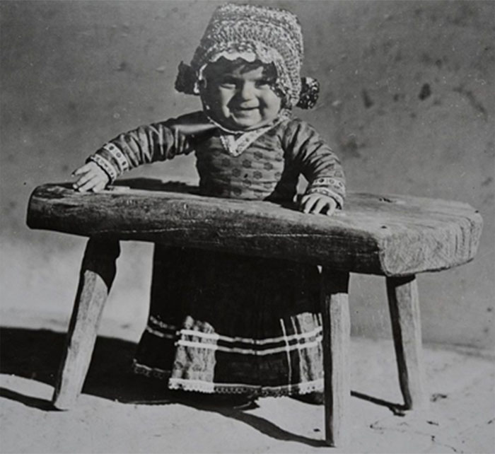 Vintage black and white photo of a child in traditional clothing standing behind a rustic wooden table, historical photo perspective.