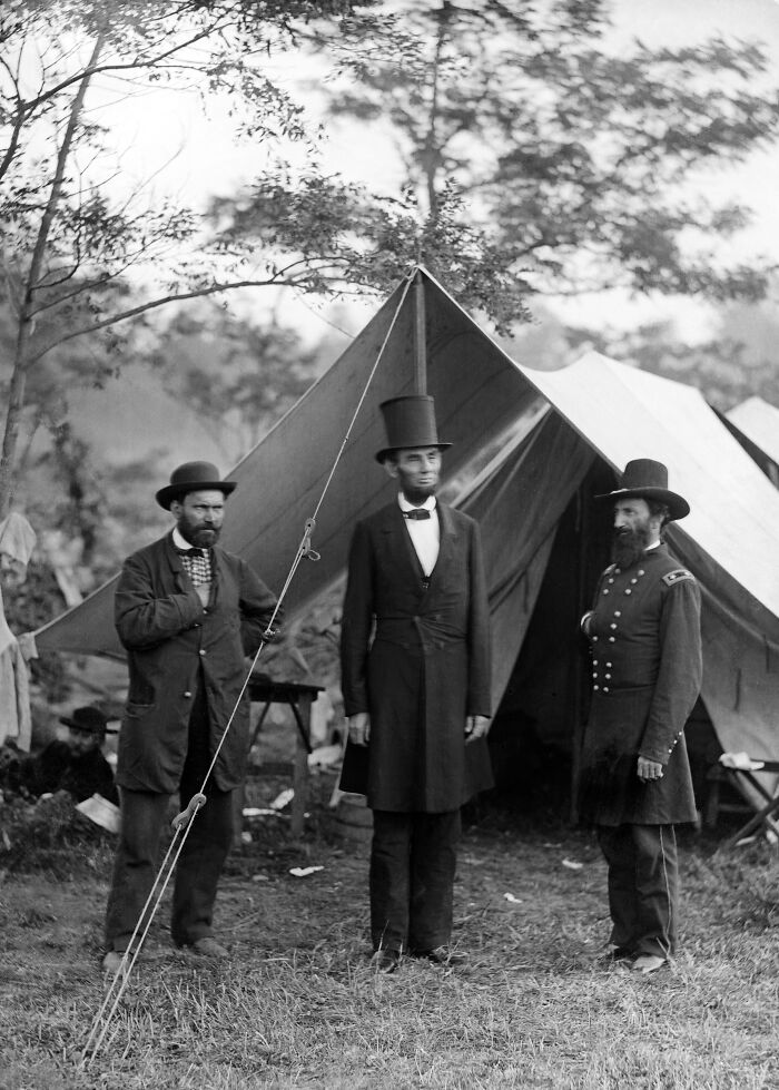 Black and white historical photo featuring men in 19th-century attire standing outside a military tent in a camp setting.