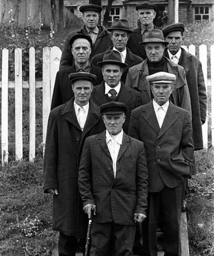 Group portrait of men in early 20th century attire standing in front of a wooden fence in a fascinating historical photo.