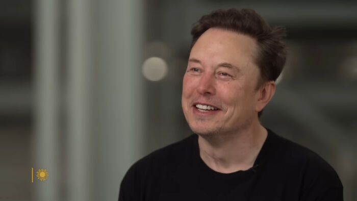 Elon Musk smiling in a casual black shirt during an indoor interview about historical folks people would love to call.