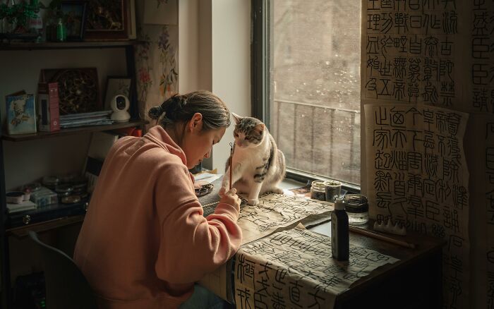 A woman practicing calligraphy with a cat beside her in a cozy room filled with historical and cultural decorations.