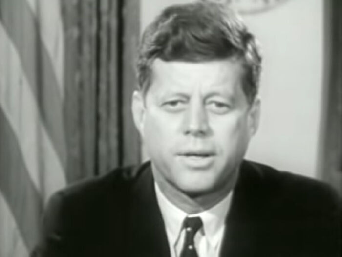 Black and white portrait of a historical figure speaking in a formal setting with a flag in the background.
