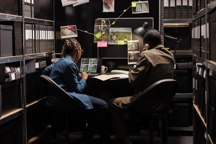 Two investigators analyzing evidence and photos in a dimly lit room filled with files, studying historical folks.
