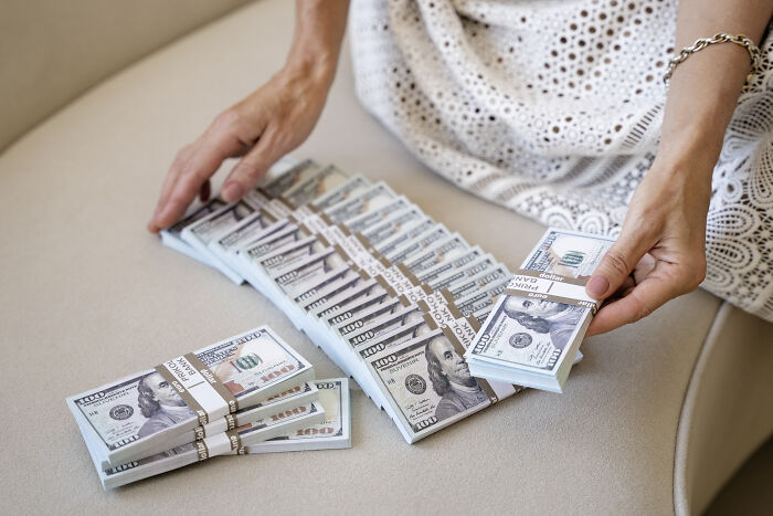 Person counting large stacks of hundred-dollar bills on a beige surface representing wealth and historical figures.