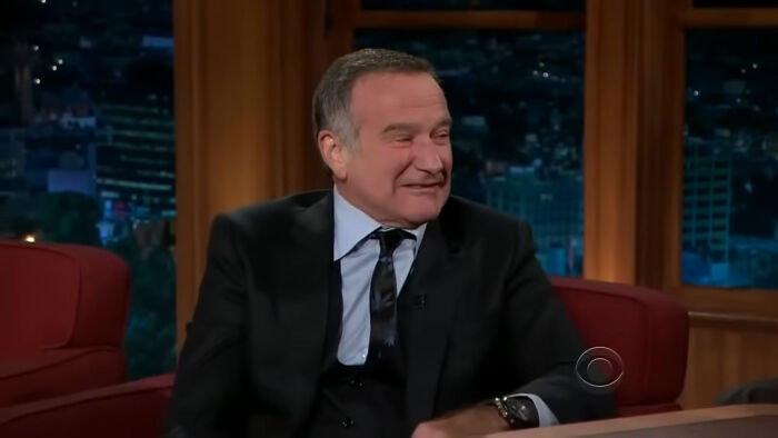 Man in a black suit and tie smiling during a late night talk show interview about famous historical figures.