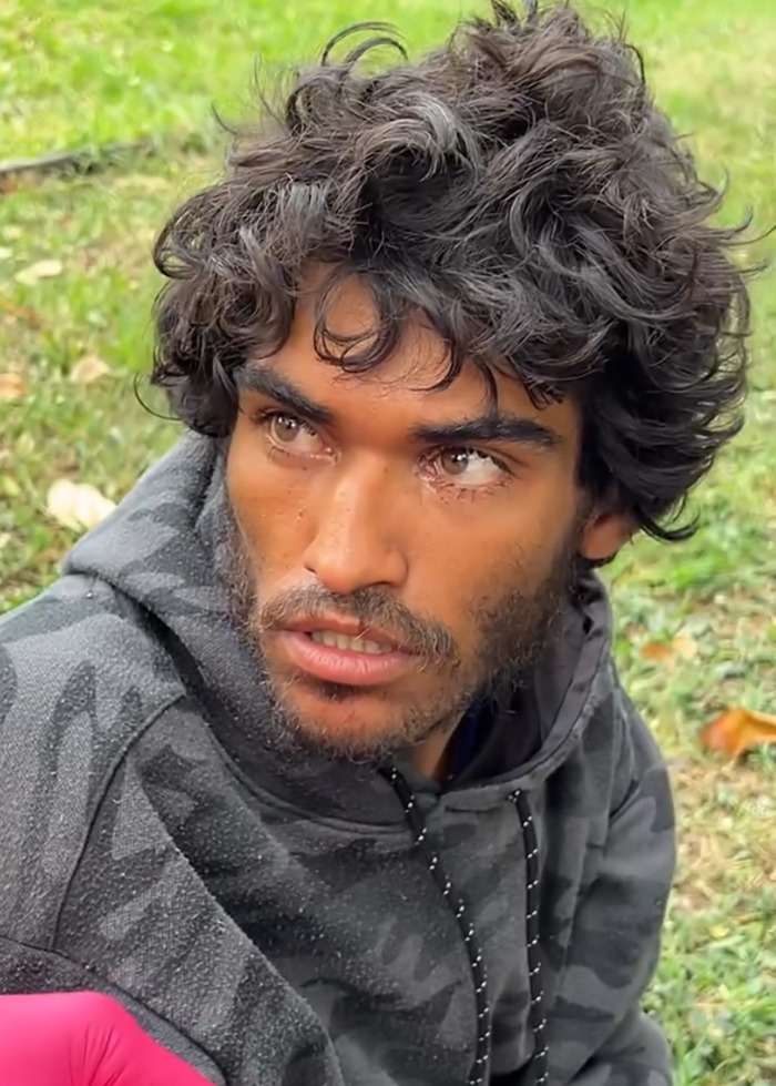 Homeless man with curly hair and beard sitting outdoors before a viral makeover and barber controversy.
