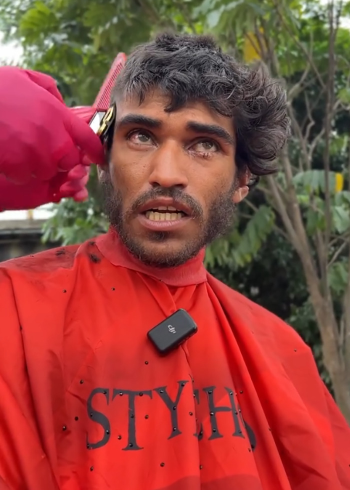 Homeless man receiving a viral makeover haircut outdoors, barber using clippers with red cape and gloves.
