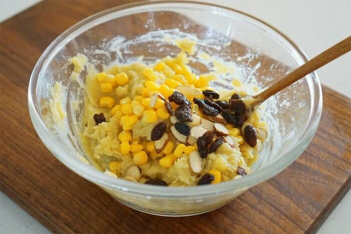 Bowl of mixed ingredients including corn, raisins, and sliced almonds being stirred with a wooden spoon on a wooden board.