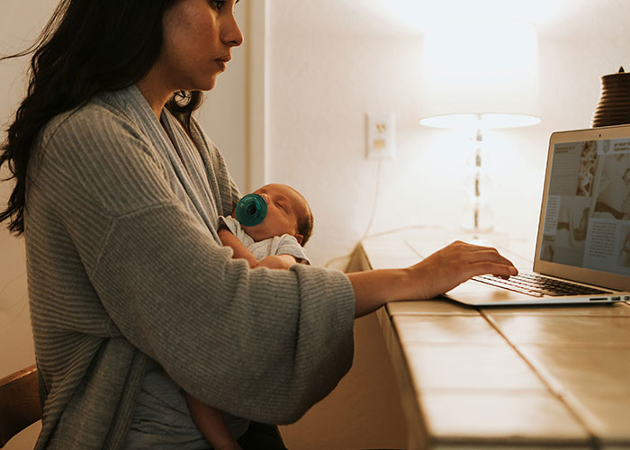Pregnant woman holding baby with pacifier, working on laptop, illustrating maternity leave eligibility dispute.